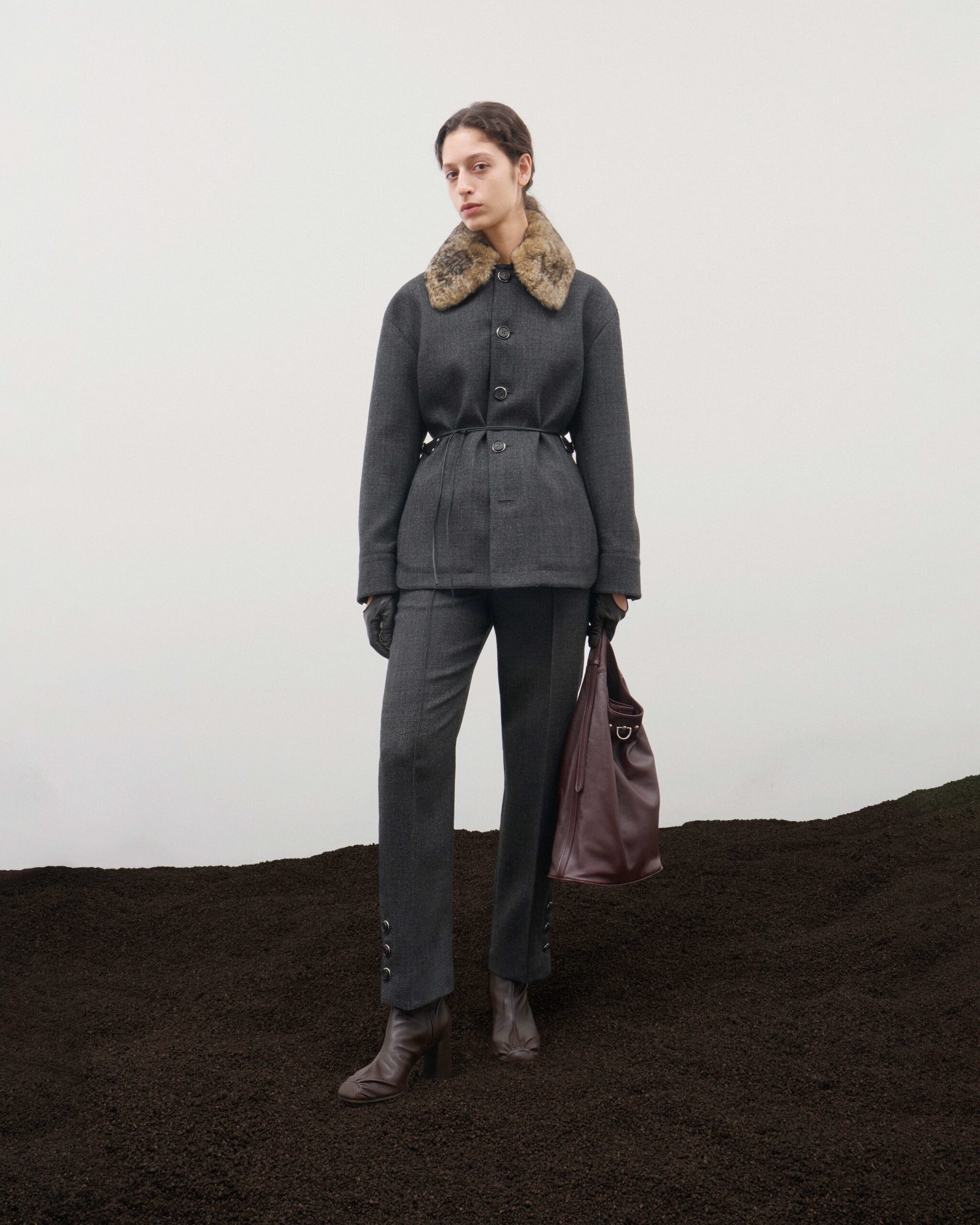 Belted grey wool jacket with fur collar over tailored trousers, burgundy leather tote bag, brown ankle boots and forest green leather gloves, Durazzi Milano FW26