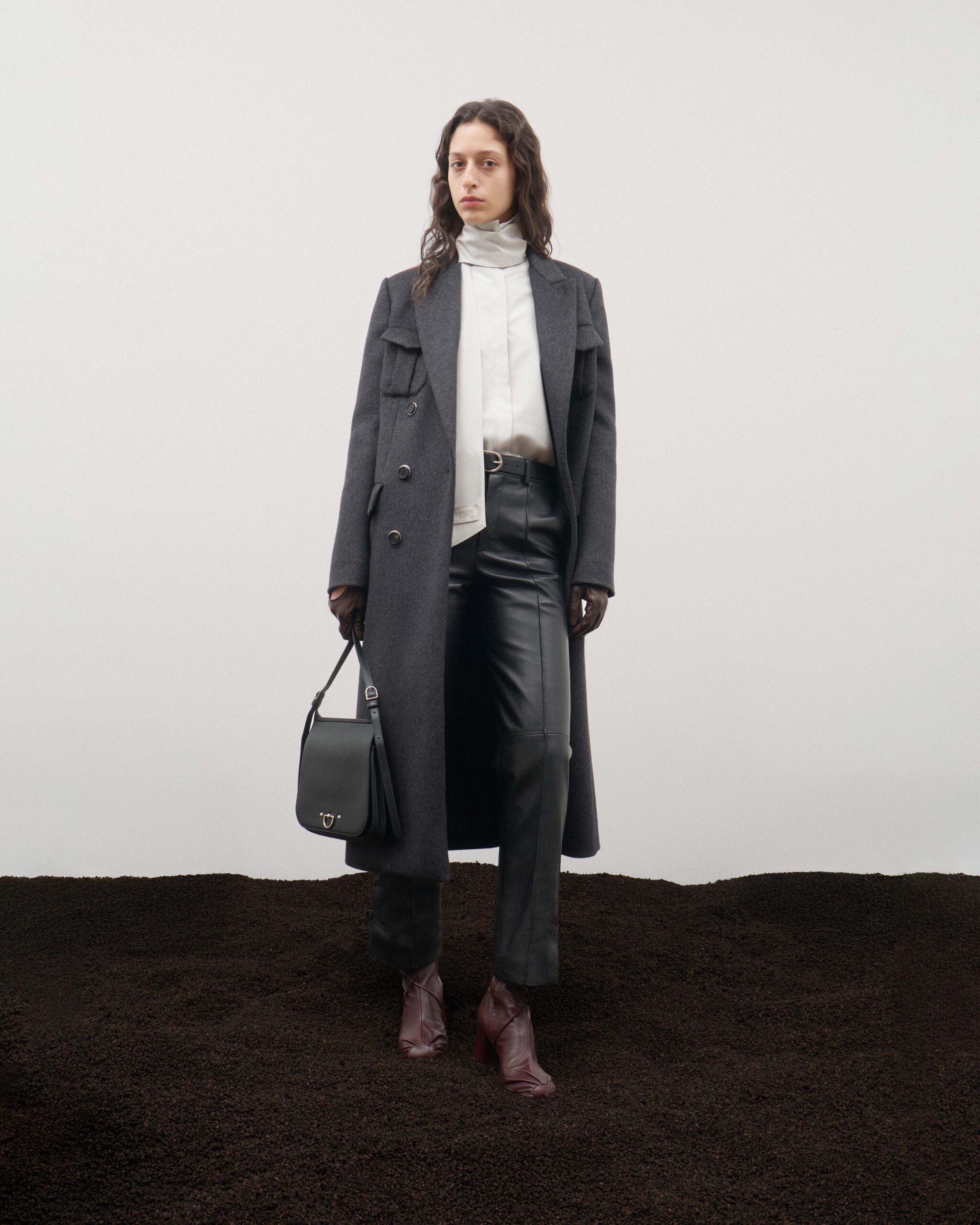 Long charcoal wool coat over ice silk shirt with removable foulard and leather trousers, brown gloves, brown ankle boots and black leather satchel, Durazzi Milano FW26