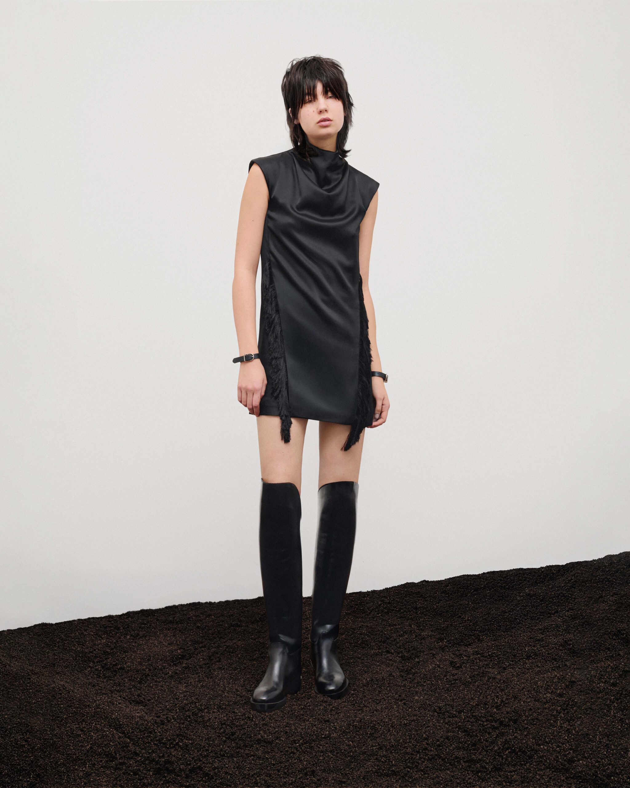 Sleeveless satin mini dress with cowl neck and fringed side worn with riding boots, Durazzi Milano FW26