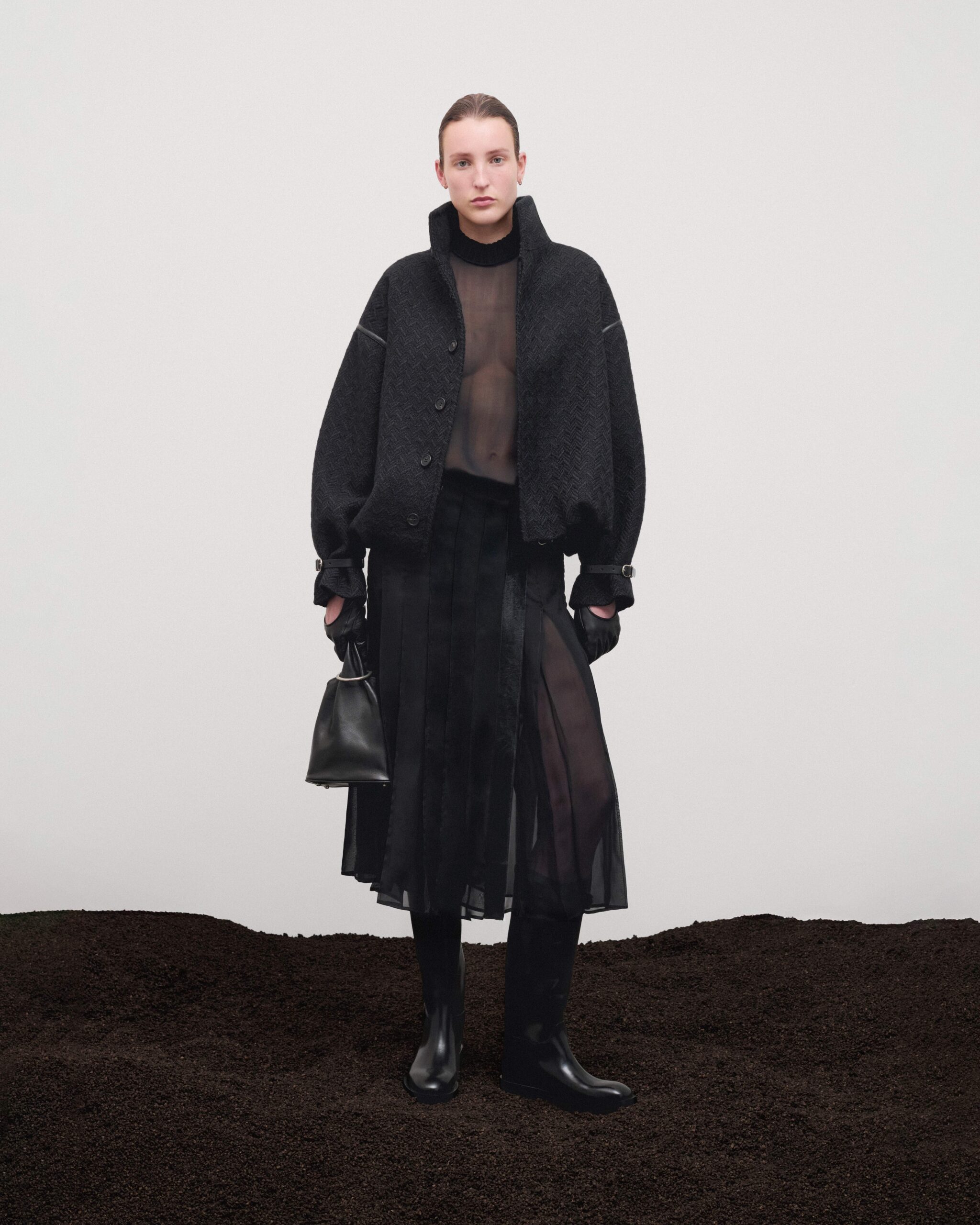 Oversized virgin wool jacket with cocoon volume over sheer top and pleated skirt, black leather gloves, black leather pouch bag and riding boots, Durazzi Milano FW26