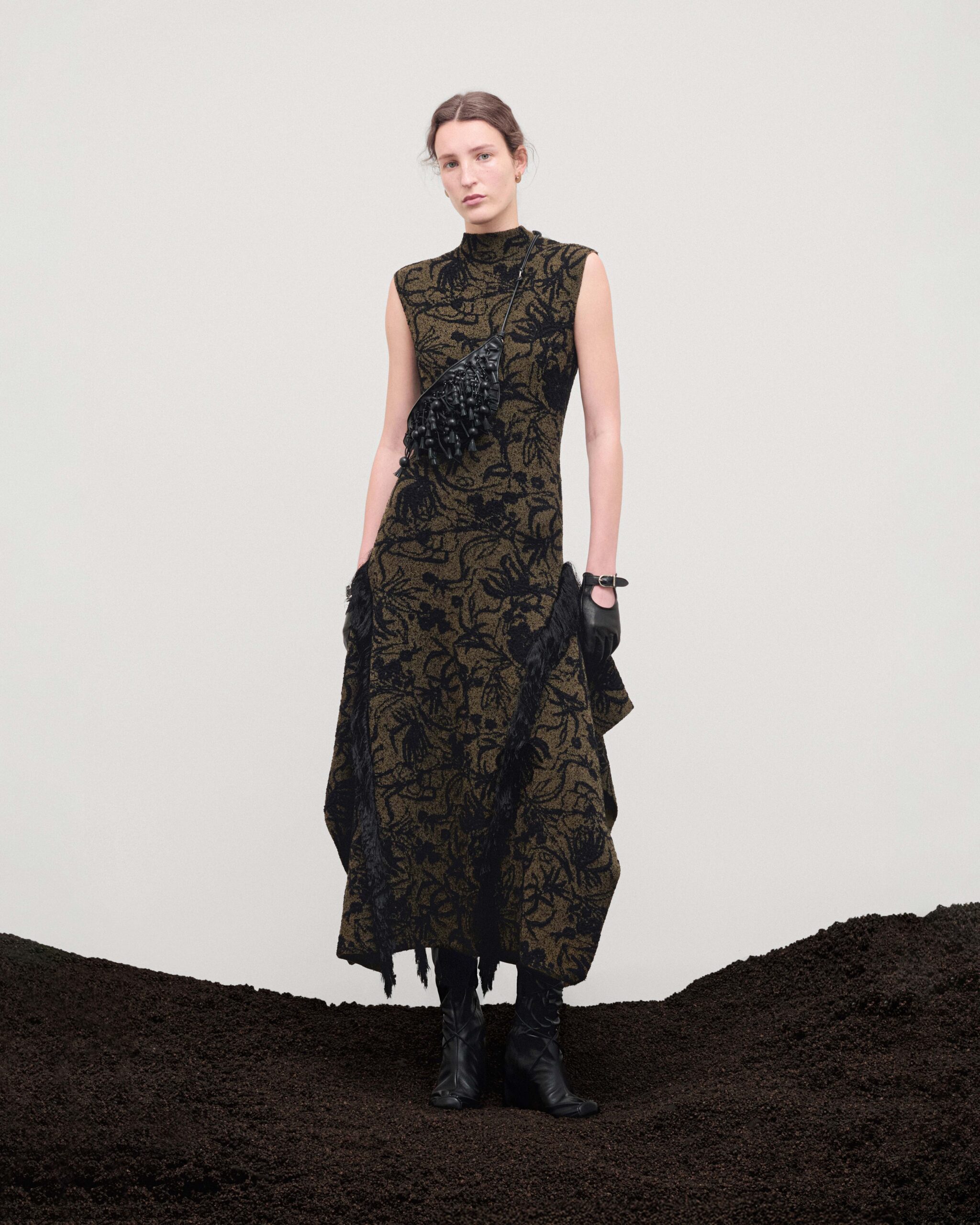Sleeveless floral jacquard long dress in olive and black with draped fringing, tasselled leather belt bag, black leather gloves and thigh-high boots, Durazzi Milano FW26